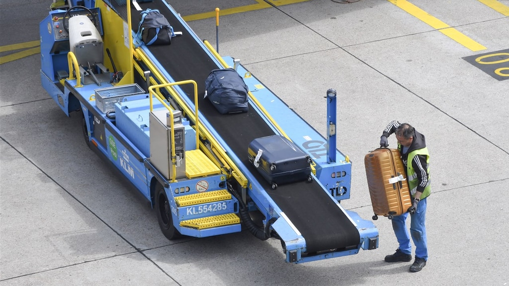Een medewerker van Schiphol tilt koffers op een band gekoppeld aan een vliegtuig.