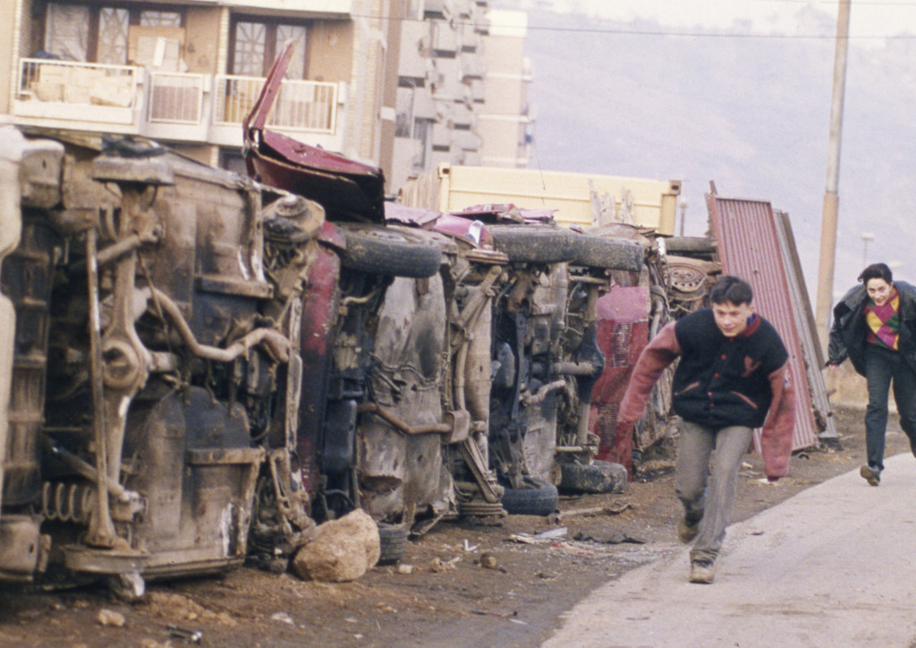 Twee Bosnische jongens zoeken dekking voor sluipschutters achter een muur die is gebouwd van autowrakken in de buitenwijk Dobrinja van Sarajevo, 1993